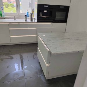 Nowoczesna kuchnia z białymi szafkami i marmurowym blatem, jasne wnętrze, duże okno z widokiem na zieleń.
